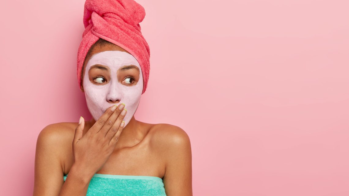 Horizontal shot of surprised African American woman covers mouth, applies nourishing mask for removing dead cells, wears wrapped towel on head, stands against pink background. Skin care concept